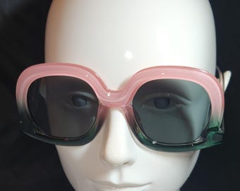 May include: Oversized sunglasses with a pink and green gradient frame and dark lenses. The sunglasses are displayed on a white mannequin head, showcasing their bold, retro design. A stylish accessory for eye protection.