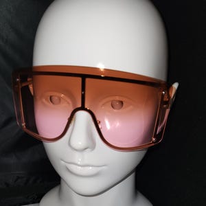 May include: A pair of oversized sunglasses with a brown to pink gradient lens. The sunglasses have a thick, square frame.