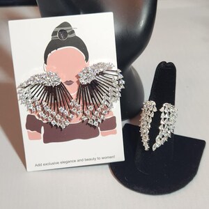 May include: A pair of silver earrings with a fan-shaped design and clear rhinestones. The earrings are displayed on a white card with a drawing of a woman wearing the earrings. The card also includes the text "Add exclusive elegance and beauty to women!"