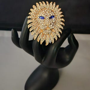 May include: A gold-toned brooch with a lion's face design. The lion's face is covered in clear rhinestones and has blue rhinestone eyes. The brooch is surrounded by a sunburst design of gold-toned metal with clear rhinestones.