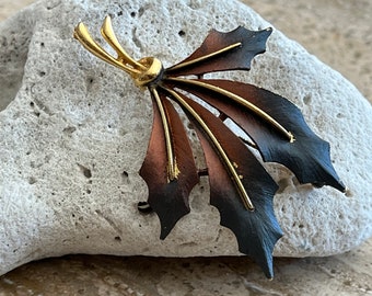 May include: A gold and copper toned leaf-shaped brooch with a textured finish. The brooch has a fan-like design with multiple layers of metal.