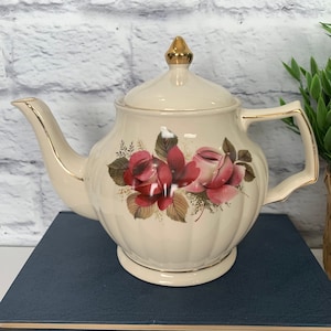 May include: Cream-coloured ceramic teapot with a lid and gold accents. The teapot features a floral design of red and pink roses. The handle and spout are trimmed with gold. The teapot is on a dark blue surface.