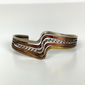 Op de afbeelding: Een metalen manchetarmband in meerdere tinten. De armband heeft lagen zilver, koper en messing in een golvend ontwerp. Een zilverkleurig kettingachtig detail is in het ontwerp verwerkt. De armband staat tegen een witte achtergrond.