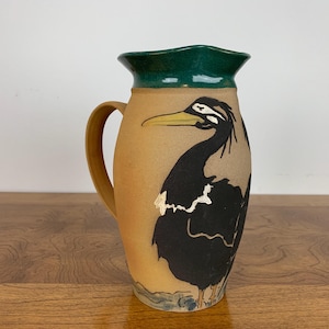 May include: A brown ceramic jug with a green lid and a black and white bird design. The bird has a long beak and is facing to the right.