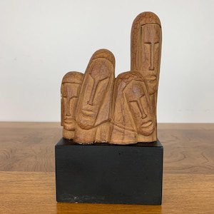 Könnte beinhalten: Eine Holzskulptur mit vier abstrakten Gesichtern, jedes mit einem einzigartigen Ausdruck. Die Skulptur ist auf einem schwarzen Holzsockel montiert.