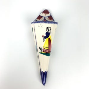 Peut inclure: Une poche murale en céramique en forme de cône, avec un motif peint à la main. La poche est principalement de couleur crème avec des accents bleus, rouges, jaunes et verts. Le motif comprend une femme en costume traditionnel, des oiseaux et des motifs floraux.