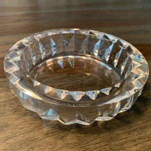 Op de afbeelding: Een glazen asbak van helder glas met een geometrisch ontwerp. De asbak is rond en heeft een verhoogde rand.