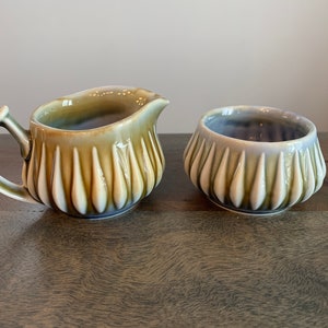 May include: A vintage Irish Belleek creamer and sugar bowl set. Both pieces are made of white porcelain with a green and brown glaze. The creamer has a handle and a spout. The sugar bowl is round and has a scalloped edge.