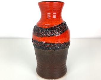 Puede incluir: Un jarrón de cerámica con una parte superior de color naranja vibrante y una banda negra texturizada alrededor del centro. La base es de color marrón oscuro. El jarrón tiene un acabado brillante y una apariencia única, hecha a mano.
