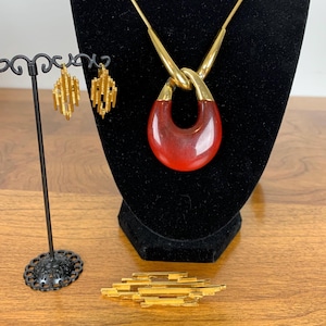 Op de afbeelding: Een gouden ketting met een rode hanger en bijpassende oorbellen. De hanger is een groot, ovaalvormig stuk rood glas met een gouden knoop aan de bovenkant. De oorbellen zijn klein en delicaat, met een geometrisch ontwerp.