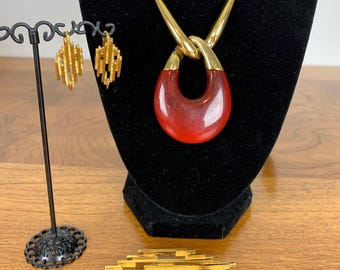 Puede incluir: Un collar de oro con un colgante rojo y pendientes a juego. El colgante es una pieza grande de vidrio rojo de forma ovalada con un nudo de oro en la parte superior. Los pendientes son pequeños y delicados, con un diseño geométrico.
