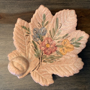 Peut inclure: Un plat en céramique en forme de feuille avec une surface texturée et un motif floral peint. Le plat présente un groupe de fleurs dans des tons de bleu, jaune, rose et vert.