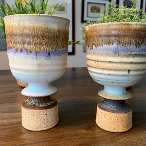 Puede incluir: Dos tazas de cerámica con esmalte marrón, blanco y azul. Las tazas tienen una base ancha y una parte superior estrecha. Están sobre una mesa de madera.