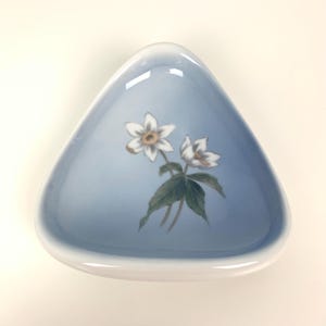 Puede incluir: Un plato triangular de cerámica azul claro con un borde blanco. El plato presenta un diseño floral de flores blancas con centros amarillos y hojas verdes. Las flores están pintadas sobre la superficie azul del plato.