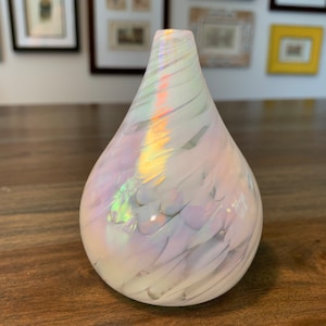 Peut inclure: Un vase en verre blanc nacré avec des tourbillons irisés. Le vase a une forme de larme et est posé sur une surface en bois.