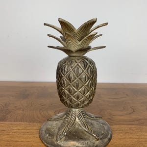 May include: A silver pineapple-shaped candle holder with a detailed design. The holder has a round base and a pineapple-shaped body with a detailed design.
