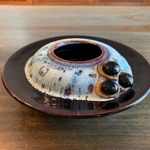 May include: A ceramic dish with a black glaze and a white and brown ring around the centre. The dish has three black round accents on the rim.
