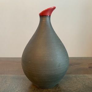Puede incluir: Un jarrón de cerámica gris oscuro con un borde rojo. El jarrón tiene una forma redondeada en forma de lágrima y una superficie texturizada.
