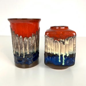 Puede incluir: Dos jarrones de cerámica con un diseño de esmalte goteado texturizado. Los jarrones presentan una combinación de colores rojo, crema, azul y marrón. Uno tiene forma cilíndrica alta y el otro una forma rectangular más corta.