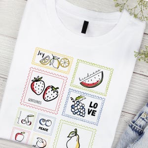 Könnte beinhalten: Weißes T-Shirt mit farbenfrohen Illustrationen von Früchten und den Wörtern "Freude", "Freundlichkeit", "Sanftmut", "Frieden" und "Liebe". Das Design umfasst Erdbeeren, Zitronen, eine Wassermelone, Trauben, Kirschen, Blaubeeren, einen Apfel und eine Birne.