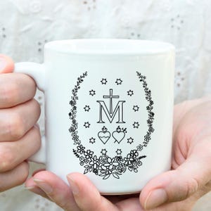 May include: White ceramic mug featuring a black floral wreath design with stars, a cross, the letter "M", and two hearts. The mug has a curved handle and is held by hands.