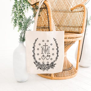May include: A cream-colored tote bag with a black floral and religious design. The design includes a cross, the letter "M", two hearts, stars, and a floral wreath. The bag has two handles and is made of a canvas-like material.
