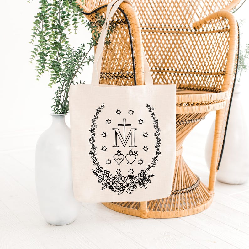 May include: A cream-colored tote bag with a black floral and religious design. The design includes a cross, the letter "M", two hearts, stars, and a floral wreath. The bag has two handles and is made of a canvas-like material.