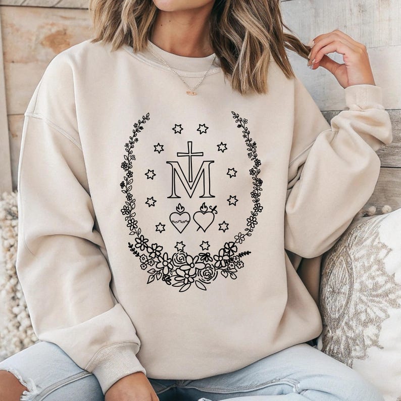 May include: Beige sweatshirt featuring a black floral wreath design with a cross, the letter "M", stars, and two hearts. The design is centered on the front of the long-sleeved, crew neck top.