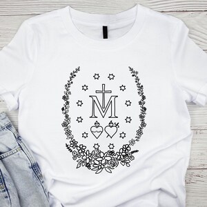 May include: White t-shirt featuring a black floral wreath design with stars, a cross, and the letter "M". Two hearts are also included in the design. The shirt is made of a soft material.