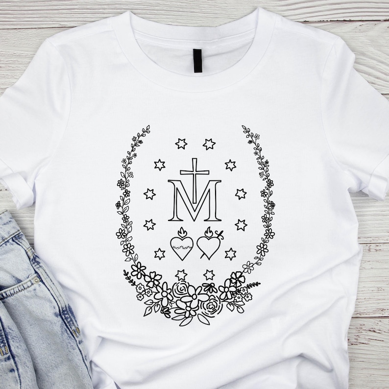 May include: White t-shirt featuring a black floral wreath design with stars, a cross, and the letter "M". Two hearts are also included in the design. The shirt is made of a soft material.
