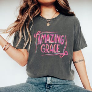 Puede incluir: Camiseta gris oscuro con las palabras "AMAZING GRACE" en una fuente colorida y estilizada. El texto está delineado en rosa y morado, con un acento azul claro. La camiseta es de estilo casual y cómodo.