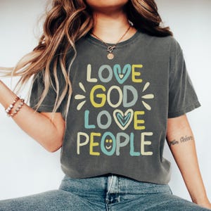 Puede incluir: Camiseta gris oscuro con las palabras "LOVE GOD LOVE PEOPLE" en una fuente colorida y juguetona. El diseño incluye gráficos de corazón y rayos de sol. La camiseta es de estilo casual y cómodo.