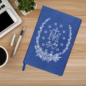 May include: A blue journal with a white floral and star design, a cross, and the letter "M". The journal is on a wooden surface with a pen, a cup of coffee, a laptop, and a small plant.