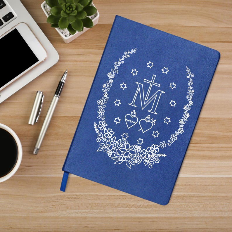 May include: A blue journal with a white floral and star design, a cross, and the letter "M". The journal is on a wooden surface with a pen, a cup of coffee, a laptop, and a small plant.
