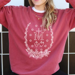 May include: A maroon sweatshirt with a white floral and star design surrounding a cross and the letter "M". The design also includes two hearts. The sweatshirt has a crew neck and long sleeves.