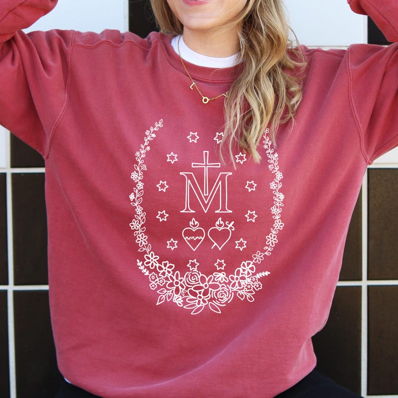 May include: A maroon sweatshirt with a white floral and star design surrounding a cross and the letter "M". The design also includes two hearts. The sweatshirt has a crew neck and long sleeves.