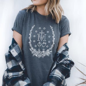 May include: Dark gray t-shirt featuring a white floral wreath design with a cross, the letter M, stars, and hearts. The shirt has short sleeves and a crew neck. The person is wearing a blue and black plaid shirt over the t-shirt.