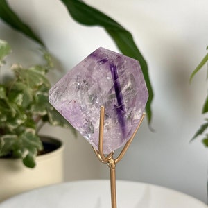 May include: A purple amethyst crystal point on a gold metal stand. The crystal is clear with purple streaks and is displayed on a white surface.