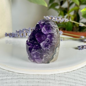 May include: A natural amethyst crystal geode, showcasing a deep purple interior with crystalline formations. The geode sits on a white plate, with lavender sprigs and greenery in the background. The geode is a unique decorative item.