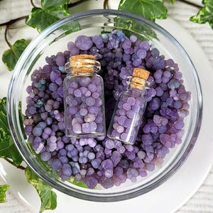 May include: Two small glass jars filled with purple agate stones. The jars have cork stoppers and are sitting in a clear glass bowl filled with more purple agate stones.
