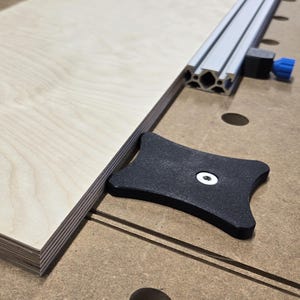 May include: A light-coloured rectangular piece of wood sits on a workbench, next to a metal guide rail and a black plastic fixture. The wood grain is visible. The workbench is brown with several holes.