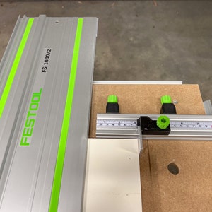 May include: A silver and green Festool guide rail with the model number FS 1080/2 printed on it. The guide rail is attached to a black and silver measuring tool with green knobs.