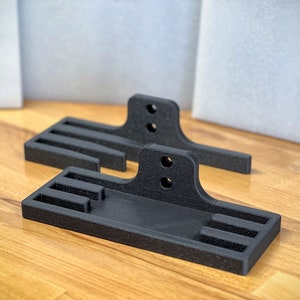 May include: Two black, rectangular, 3D-printed objects with a flat base, vertical supports, and two holes. The objects have slots and are on a wooden surface. The background includes white and gray elements.