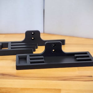 Puede incluir: Dos soportes de pared de plástico negro con ranuras para sujetar objetos. Los soportes tienen dos agujeros en la parte posterior para sujetarlos a una pared.