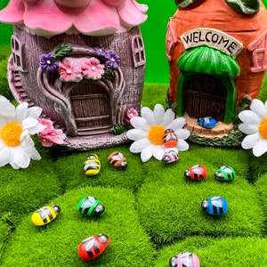 May include: Two miniature fairy houses, one pink and one orange, with a welcome sign, surrounded by green grass and colorful wooden bee figurines.