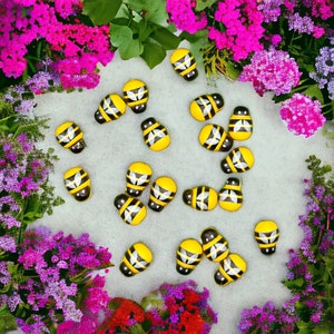 May include: A collection of yellow and black bee-shaped wooden ornaments on a white background surrounded by pink and purple flowers.
