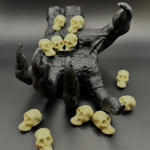 May include: A black skeletal hand sculpture with a hollowed-out palm, holding a collection of small, off-white skull figurines. Several more skulls are scattered around the base of the hand.