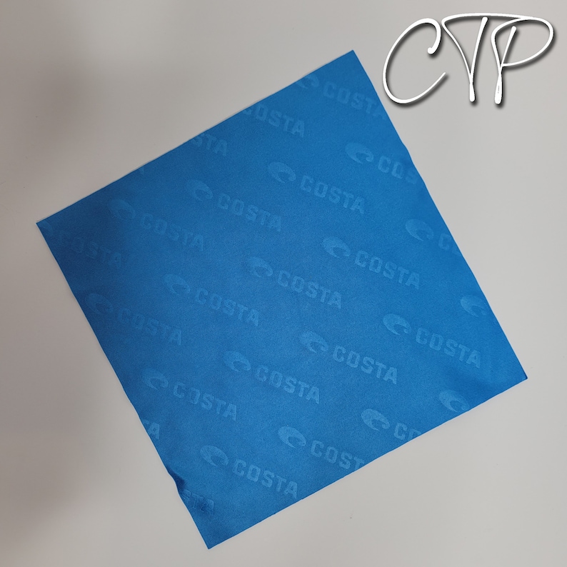 May include: A square, bright blue cloth with the word "COSTA" repeatedly printed across the surface. The logo is a stylized "C" shape. The cloth is slightly wrinkled and appears to be made of a soft material. The letters "CYP" are in the upper right corner.
