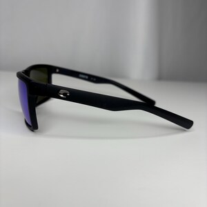 May include: Black sunglasses with a matte finish and blue-tinted lenses. The sunglasses have a rectangular frame and a small silver logo on the temple. The arms are thin and curve slightly.