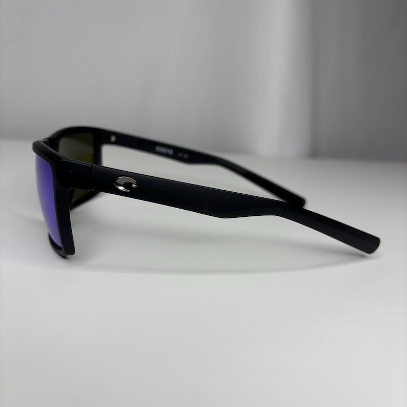 May include: Black sunglasses with a matte finish and blue-tinted lenses. The sunglasses have a rectangular frame and a small silver logo on the temple. The arms are thin and curve slightly.